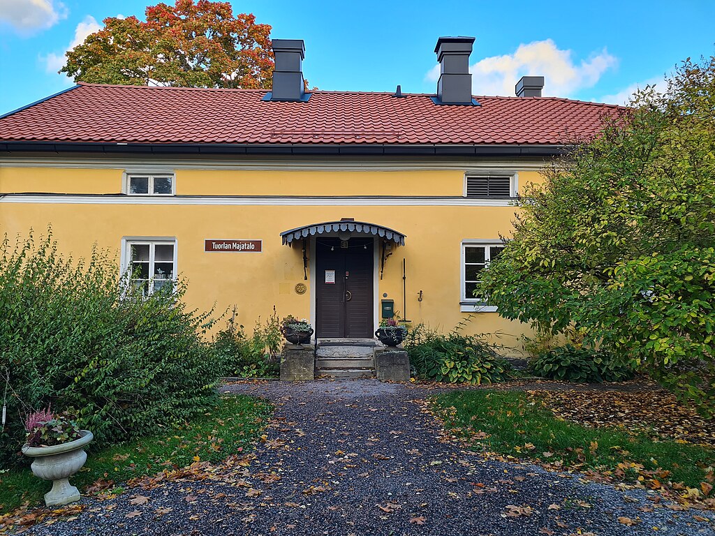 Kuvassa on sateinen syksyinen päivä Tuorlan Majatalolla. Rakennuksen seinät ovat kirkkaan keltaiset ja ovi tummanruskea. Pihaa koristavat ruukut ja kauniit syyskukat.