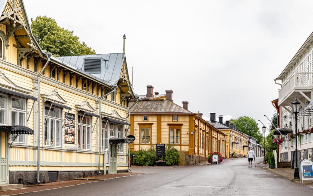 Kuva Naantalin idyllisestä kadusta, jota reunustavat vaaleat, pääosin keltaiset ja valkoiset puutalot koristeellisilla yksityiskohdilla. Kadulla kävelee yksi henkilö poispäin kamerasta, ja taloissa näkyy liikemerkkien kylttejä. Taivas on harmaa ja luo rauhallisen tunnelman.