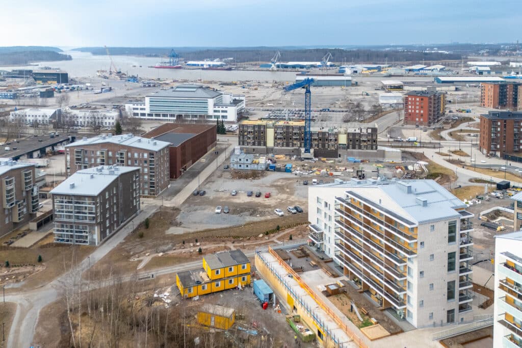 Ilmakuva rakenteilla olevasta asuinalueesta meren rannalla. Kuvassa näkyy useita kerrostaloja eri rakennusvaiheissa, nosturi keskellä työmaata sekä keskeneräisiä teitä ja pihoja. Etualalla on vaaleita ja tummia kerrostaloja sekä pieni keltainen rakennus. Taustalla avautuu satama-alue ja meri, ja ympäristö näyttää osin vielä keskeneräiseltä.